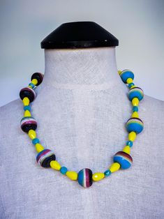 This bold oversized beaded necklace features 23mm vintage 1980s African sand cast recycled multicolor glass beads, showcasing vibrant layers of black, red, white, yellow, and blue. Complementing these striking beads are 9mm electroplated teal/blue faceted crystal coin beads, framed by 15x10mm faceted bright yellow teardrop crystal glass beads, adding an eye-catching pop of color. The collar-length design measures approximately 23 to 24.5 inches, making it a standout statement piece. The beaded s Retro Multicolor Necklaces For Jewelry Making, Retro Multicolor Round Bead Jewelry, Retro Multicolor Round Beads Jewelry, Retro Multicolor Round Beads Necklace, Retro Multicolor Round Beaded Jewelry, Retro Multicolor Beaded Necklace, Retro Multicolor Handmade Beaded Necklaces, Retro Multicolor Jewelry For Beach, Retro Multicolor Beach Jewelry