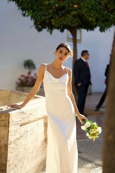 a woman in a white dress is holding a bouquet and standing next to a stone wall