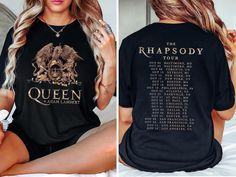 a woman sitting on top of a bed wearing a black t - shirt with the queen tour dates printed on it