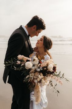 a bride and groom standing on the beach with flowers in their hands, looking at each other