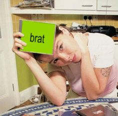 a woman holding up a sign with the word brat on it in front of her face