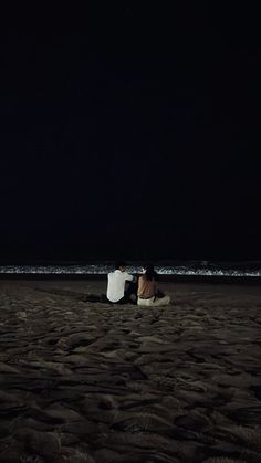 two people sitting on the beach at night with their backs to each other, facing the ocean