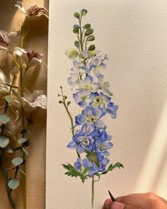 a person is holding a pencil and drawing flowers on paper with watercolors in front of them