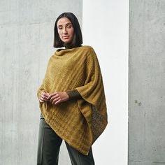 a woman standing in front of a concrete wall wearing a poncho and green pants