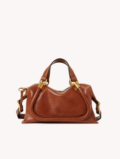 a brown handbag with gold handles and straps on the handle, sitting against a white background