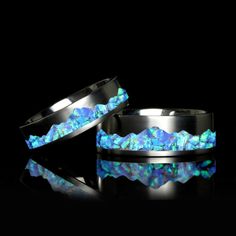 two rings with blue and green stones in them on a reflective surface, against a black background