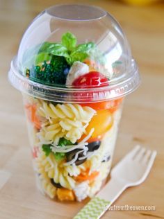 a plastic container filled with pasta and veggies on top of a wooden table