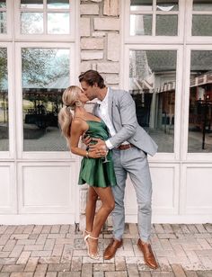 a man and woman are kissing in front of a building with large windows, one is wearing a green dress while the other wears a gray suit