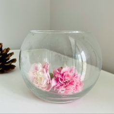 two pink flowers in a clear bowl on a white table next to a pine cone