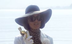 a woman in a hat and sunglasses holding an oscar statue while standing next to the ocean