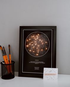 a black frame with some pens and pencils in it next to a card on a table