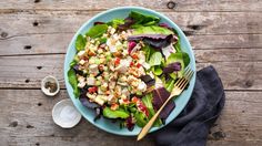 a blue bowl filled with salad next to a fork and napkin on top of a wooden table