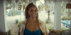 a woman in a blue top and trench coat standing on a porch with trees behind her