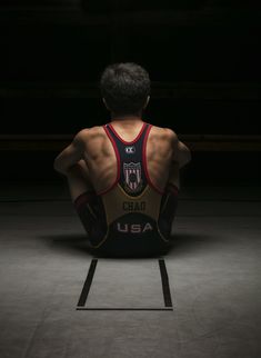 a man sitting in the dark with his back turned to the camera, wearing a wrestling uniform