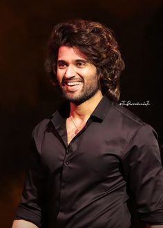 a man with long hair smiling and wearing a black shirt in front of a dark background