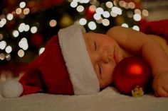 a baby wearing a santa hat sleeping next to a christmas tree with lights in the background