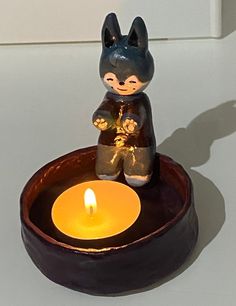 a small cat figurine sitting on top of a bowl with a lit candle