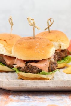 two hamburgers with toothpicks on them sitting on a wooden tray next to potato chips