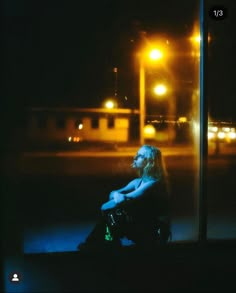 a woman sitting on the ground in front of a building at night with street lights