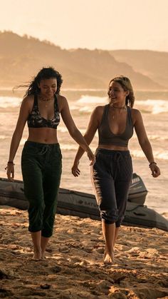 two young women walking on the beach with their hands in each other's pockets