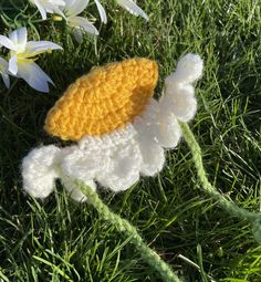 a crocheted white and yellow flower laying on top of green grass with daisies in the background