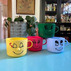 four coffee mugs with faces painted on them sitting on a table in front of a potted plant