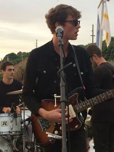 a man playing guitar and singing into a microphone at an outdoor music festival with other people