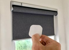 a hand is holding up a small white object in front of a window with black roller shades