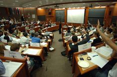 a class room full of students raising their hands in the air with papers on desks