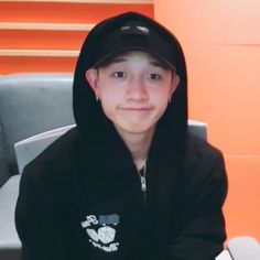 a young man wearing a black hoodie sitting in front of a gray chair with an orange wall behind him