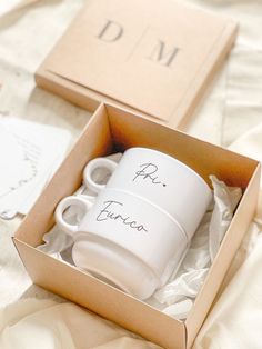 two white coffee mugs sitting in a box on top of a bed next to each other