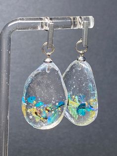 These handcrafted earrings are a true show to the artistry and creativity of their maker. Each earring features a clear resin teardrop pendant that cradles a pieces of iridescent abalone shell. The shell's natural hues ripple and shimmer within the resin, creating an enchanting play of colors that evoke the depths of the ocean. This fusion of organic beauty with the clarity of resin results in a mesmerizing and wholly unique piece of jewelry. Complementing the abalone's aquatic allure are delica Unique Iridescent Teardrop Earrings, Clear Long Drop Earrings For Gift, Resin Teardrop Earrings For Gifts, Unique Drop Earrings With Natural Inclusions, Unique Teardrop Resin Earrings, Clear Teardrop Earrings, Bar Post, Gold Flake, Silver Bar