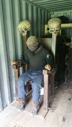 a man sitting on top of a wooden chair next to two large metal skulls hanging from the ceiling