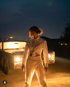 a woman standing in front of a car on the road at night with her hands behind her back