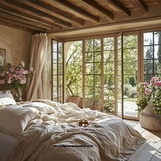 an unmade bed with white linens and pink flowers in front of large windows