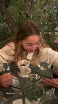 a woman sitting at a table with two wine glasses in front of her and another person holding a cup