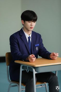 a young man in a suit sitting at a desk with his hands clasped to his knees