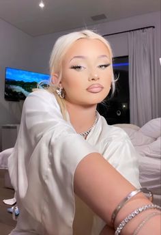 a woman with blonde hair wearing jewelry and sitting on a bed in front of a tv