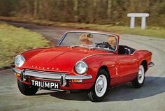 an old red triumph sports car driving down a road with two people in the back seat