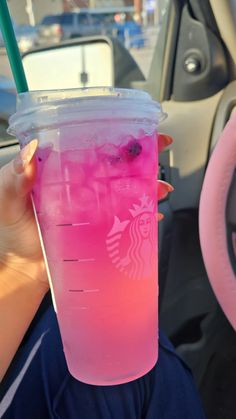 a person holding up a pink drink in their hand while sitting in a car with the steering wheel behind them