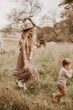 a woman in a dress and hat walking through a field next to a small boy