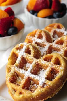 two waffles on a white plate with fruit in the background and powdered sugar on top