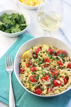 a bowl of pasta with tomatoes, parsley and olives next to a glass of white wine