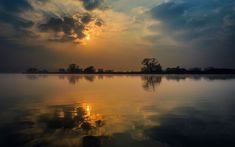 the sun is setting over some water with clouds in the sky and trees on the other side