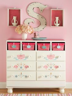 a white dresser with pink drawers and flowers on it's top, in front of a pink wall