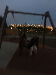 two people are playing on a swing set at night, with lights in the background