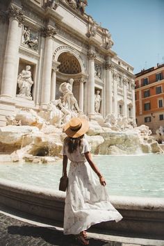 a woman in a white dress and straw hat standing near a fountain with statues on it