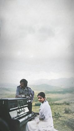 a man and woman sitting on the back of a black piano in an open field