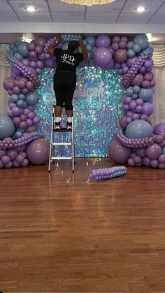 a woman standing on a ladder in front of a backdrop with purple and blue balloons