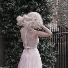 a woman with blonde hair standing in front of a fence and ivy covered wall, wearing a white dress